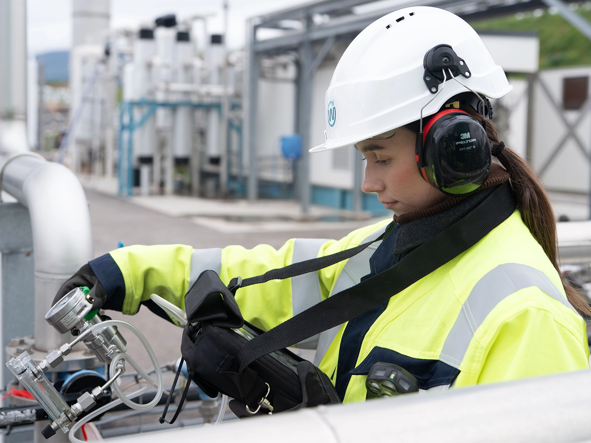Photograph of a technician wearing high‑visibility safety clothing, a helmet, and hearing protection while connecting and reading measurement instruments attached to piping at an outdoor biogas facility. Industrial structures and gas processing units are visible in the background.