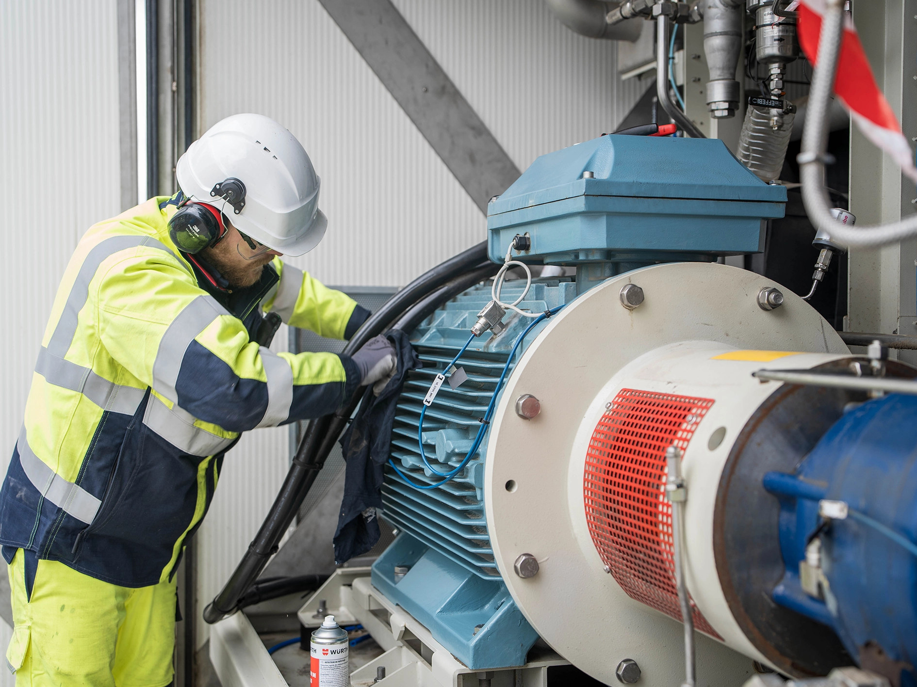 Maintenance Operation on Biogas Processing Equipment Photograph showing a technician wearing high‑visibility protective clothing, a helmet, and hearing protection while adjusting cables and components on a large industrial motor connected to gas processing equipment. Pipes, wiring, and mechanical parts are visible in a compact industrial setting.