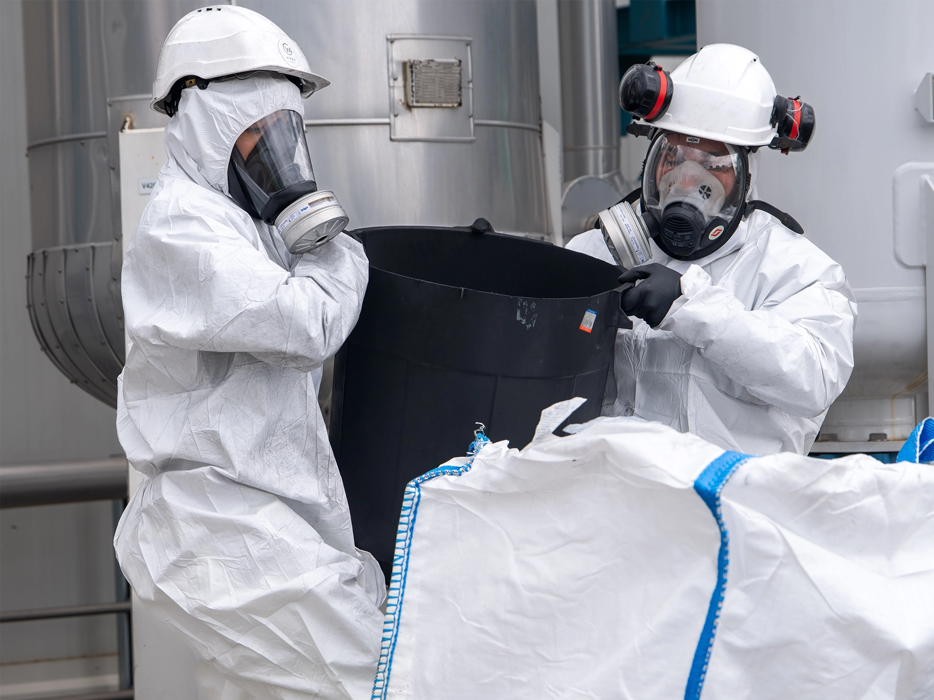 Technicians Handling Materials with Full Protective Equipment Photograph showing two technicians wearing full protective suits, helmets, and respirator masks while handling a large container or bag in an industrial installation. Large metallic tanks and piping systems are visible in the background, indicating a controlled environment operation.
