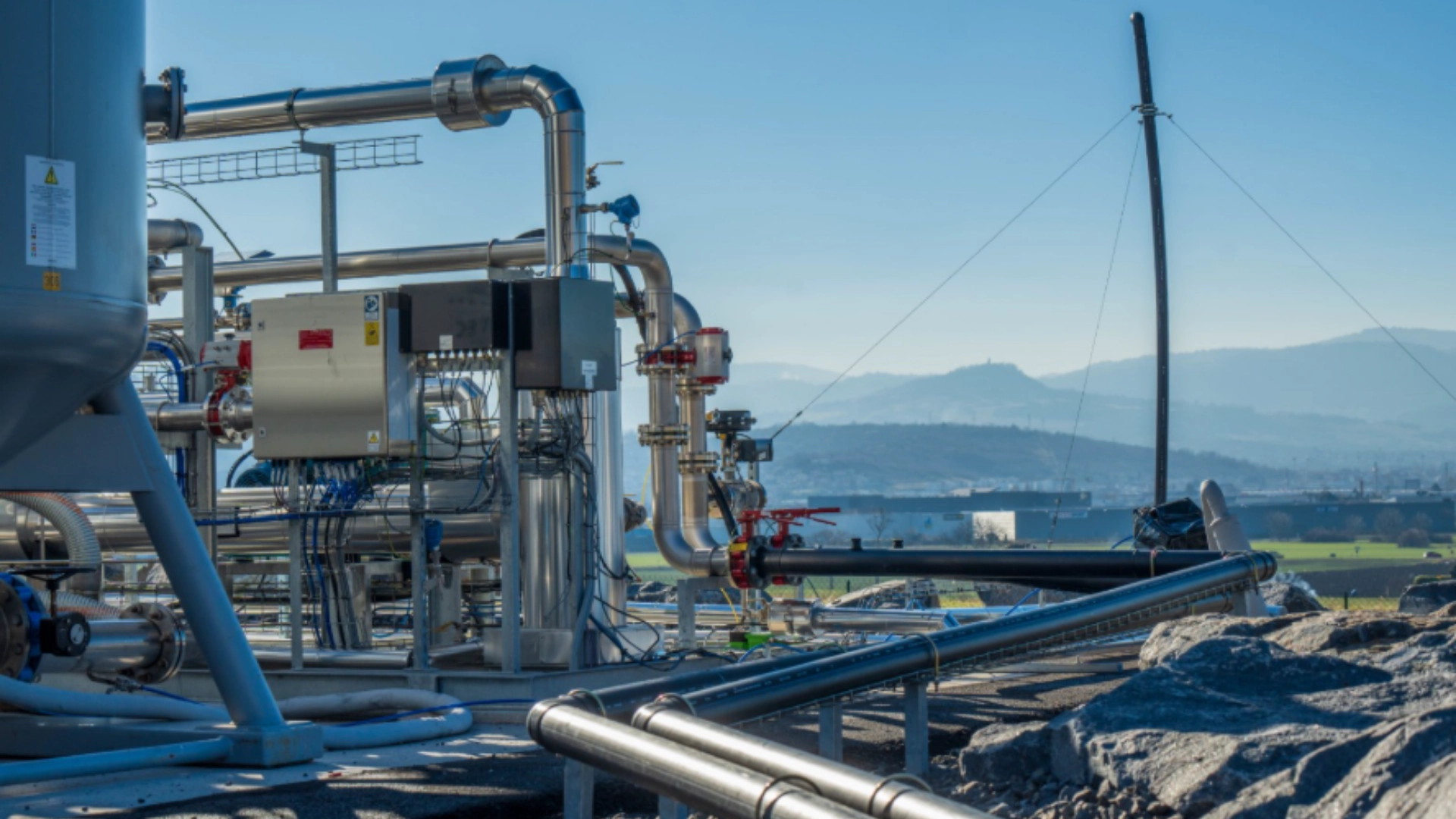 Photograph of an outdoor biogas upgrading installation featuring stainless steel pipes, valves, control panels, and processing units mounted on metal structures. The facility is located in a rural landscape with hills and fields visible in the background under clear daylight, illustrating industrial equipment used to purify biogas into renewable natural gas.