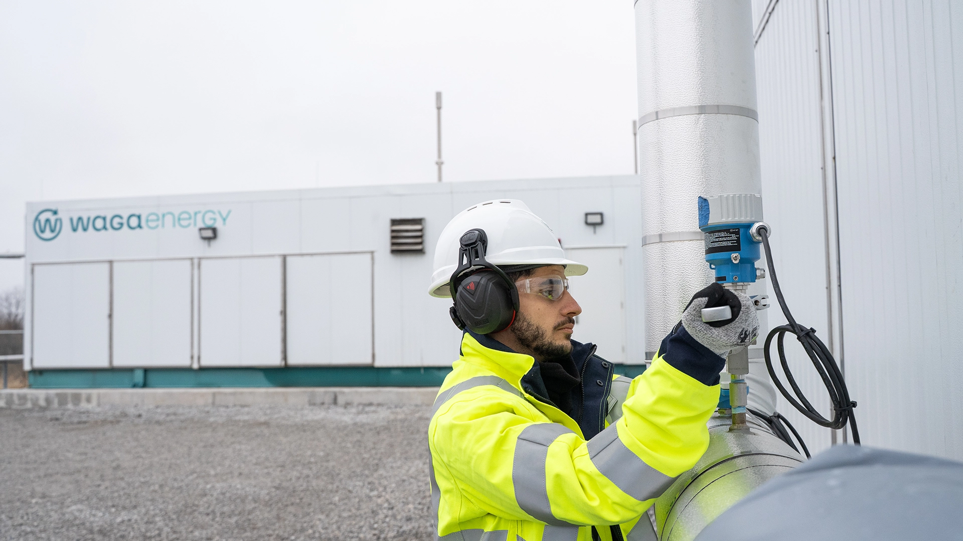 Photograph showing a worker wearing high‑visibility protective clothing, a helmet, and hearing protection while adjusting a sensor or valve on a metal gas pipe. The scene takes place outdoors at an industrial biogas upgrading facility, with processing buildings and equipment visible in the background under overcast daylight.